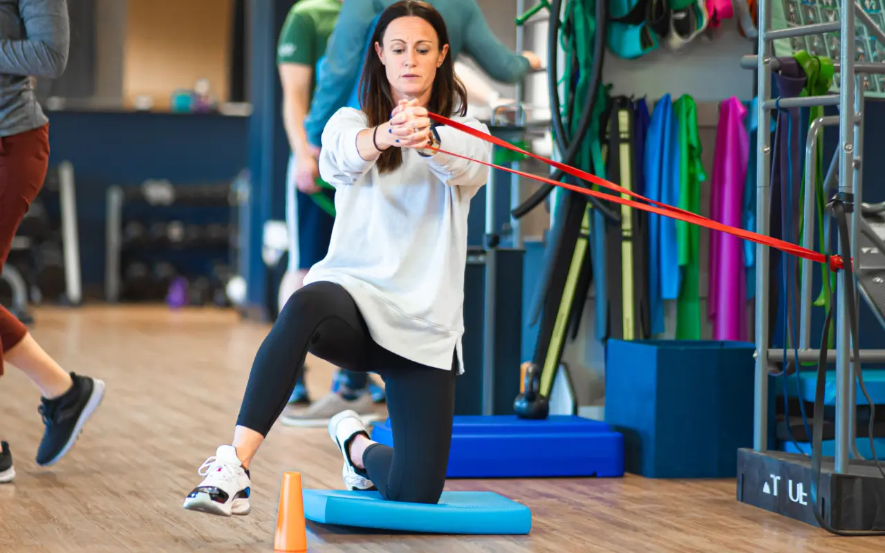 A woman kneeling on a blue pad as she lifts her leg over a cone.