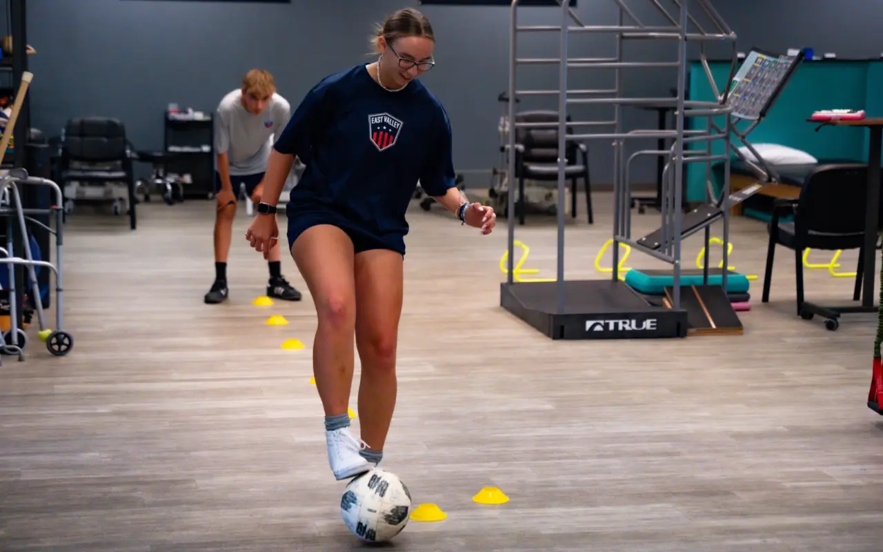 A teen athlete kicking a soccer ball around cones inside a clinic.