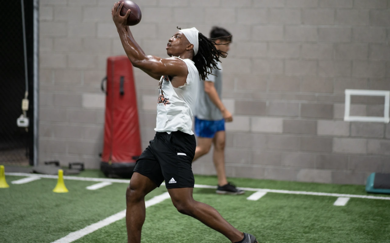 A male college athlete catches a football on an indoor turf.