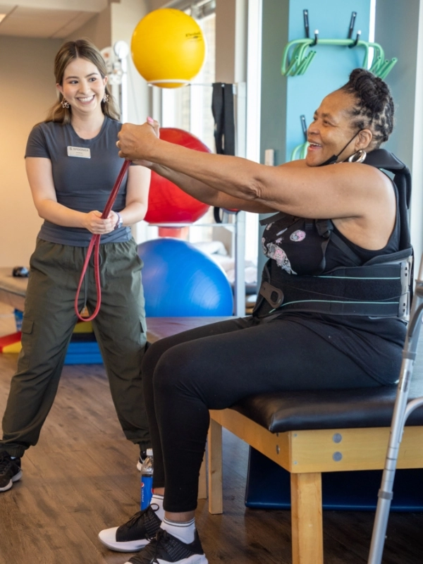 out-Spine Image 2 Image of a woman in a back brace sitting on a table with her arms outstretched forward.