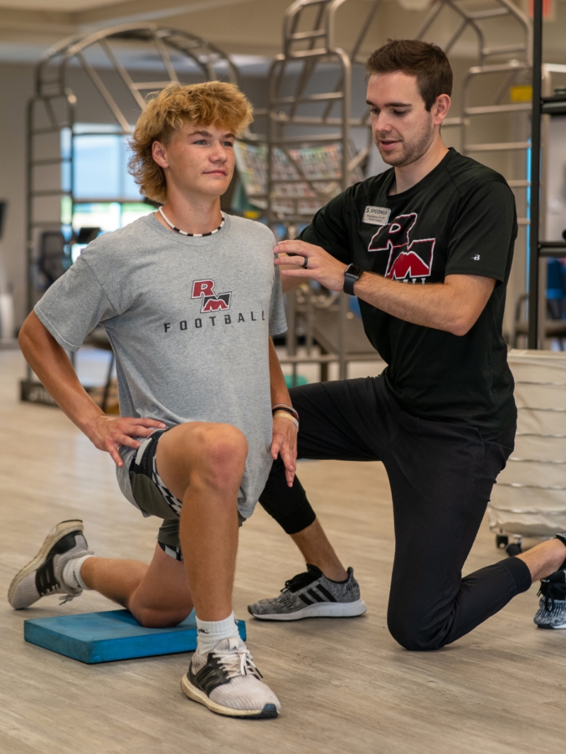 Male teen athlete kneeling on a blue pad demonstrating the frontal plane of motion by leaning forward.