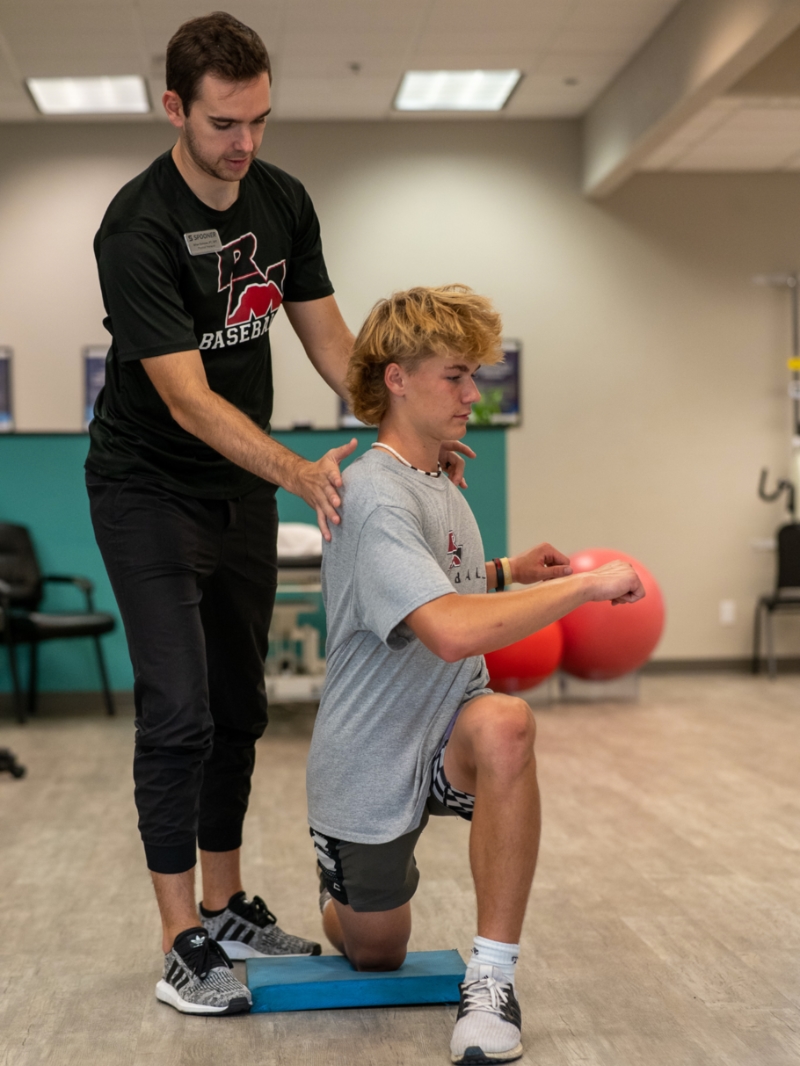 Male teen athlete kneeling on a blue pad demonstrating the transverse plane of motion by twisting the torso.