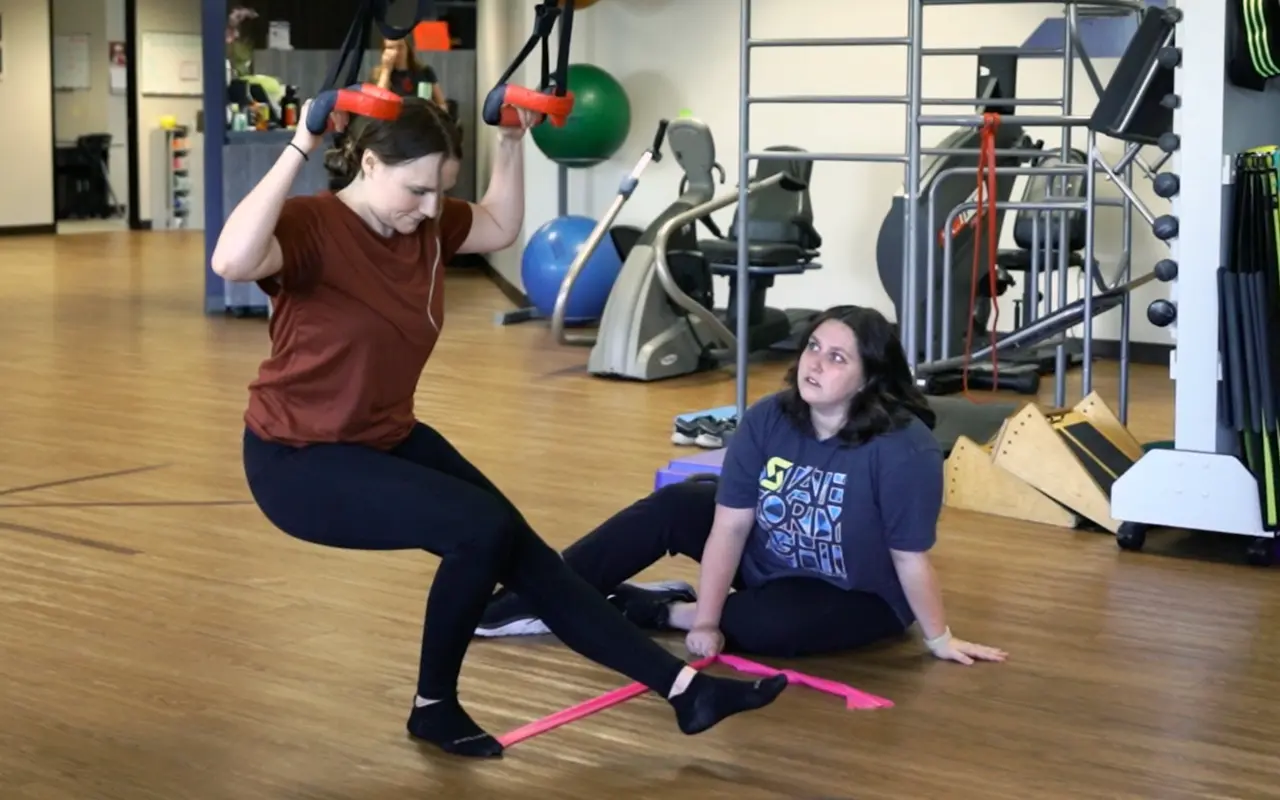A woman doing a single leg squat with TRX assistance.