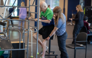 An elderly man using a stretch cage as a therapist assists him.