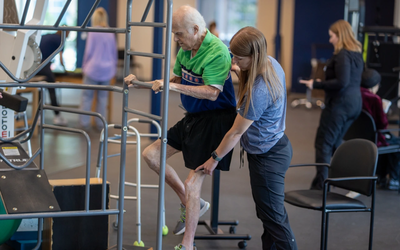 An elderly man using a stretch cage as a therapist assists him.