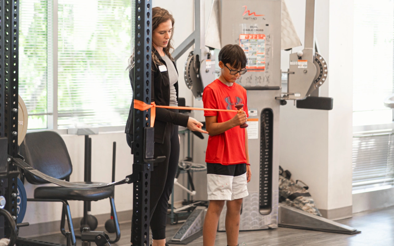 A young boy holding a dumbbell and doing elbow rotations with a resistance band.