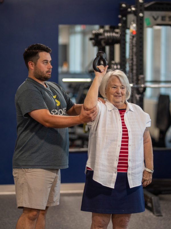A therapist steadying the arm of an older woman holding a kettlebell over her head.