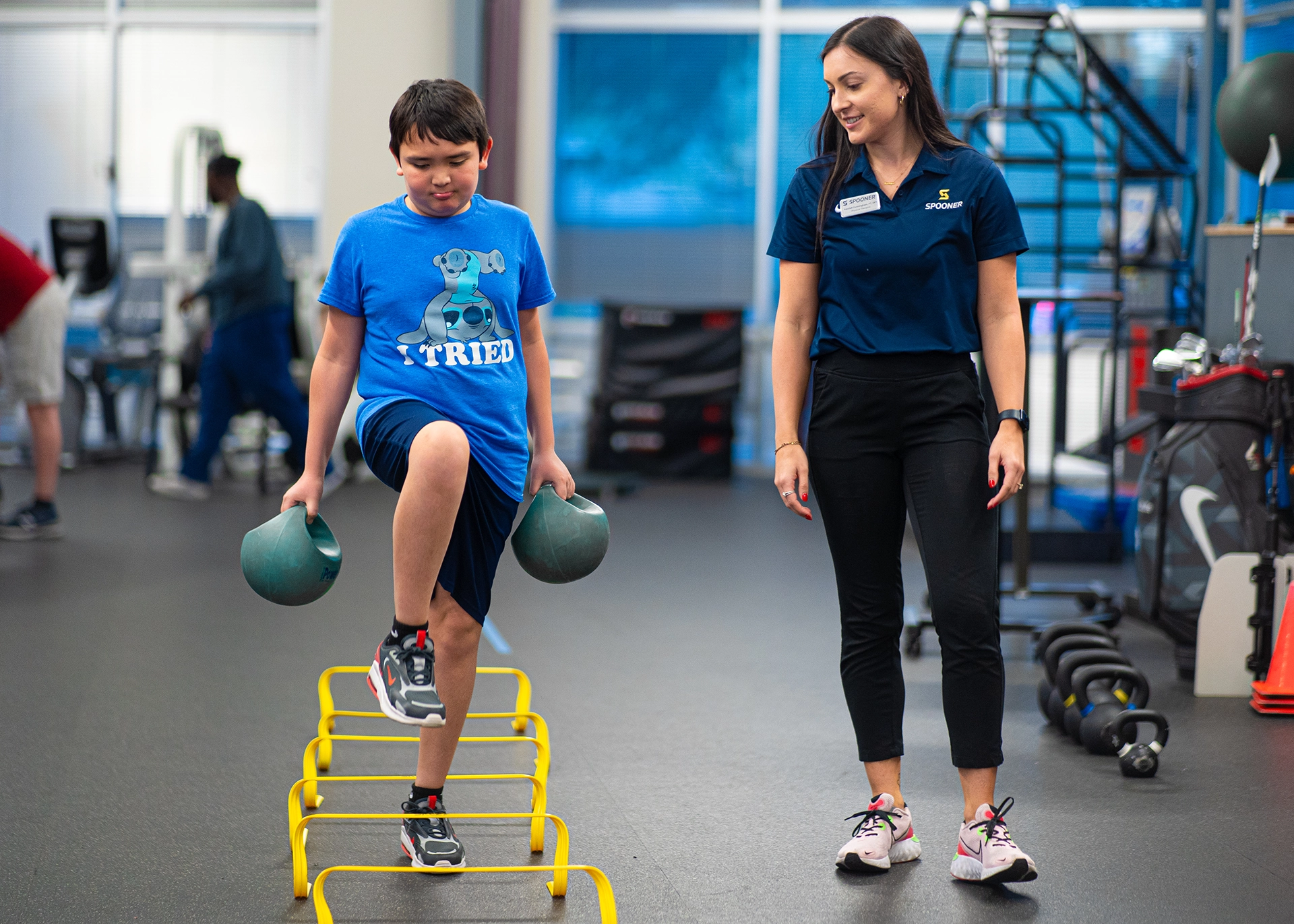 A young boy hold 2 kettlebells as he walks over low hurdles and a physical therapist monitors his balance.