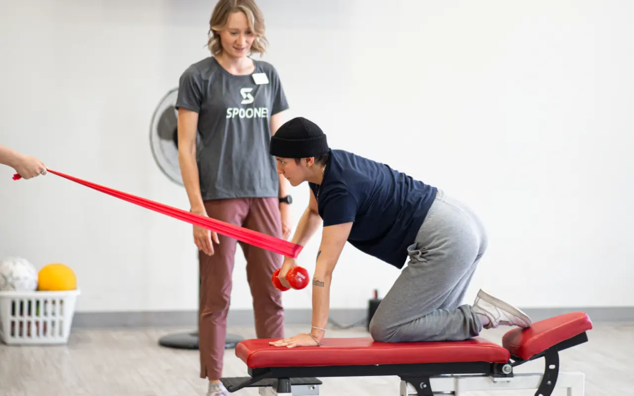 A young man on a red bench holding up a red dumbbell and a person off-screen pulls on a resistance band wrapped around his wrist.