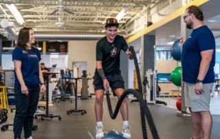 A baseball athlete swinging battle ropes as a physical therapist and a tech monitor his movement.