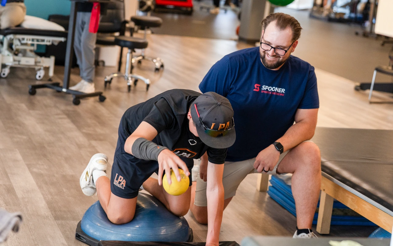A young athlete lifts a yellow ball while balancing on a bosu ball next to his physical therapist.
