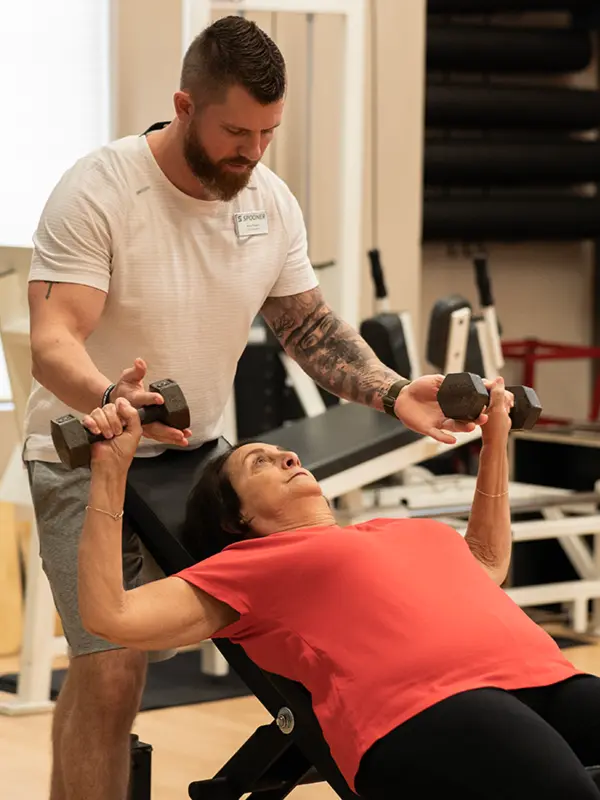 Image of a FIT Trainer spotting an older woman as she does dumbbell presses.