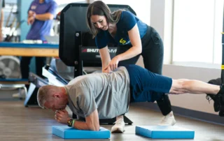 A man planking on a blue foam pad with his feet suspended in a TRX strap as a physical therapist touches the small of his back.
