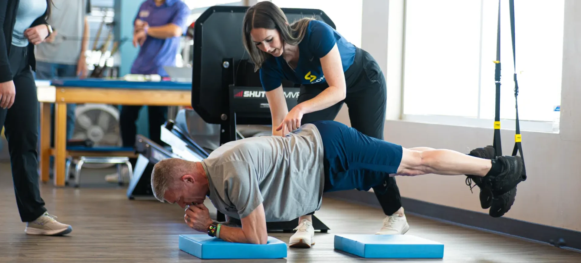 A man planking on a blue foam pad with his feet suspended in a TRX strap as a physical therapist touches the small of his back.