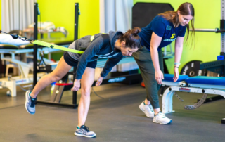 A woman stretching her hamstring with a resistance strap alongside a physical therapist.