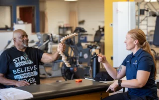 Tara Wright, OTR/L, CHT giving a man instruction on what to do with the tool he is holding for his hand exercises.