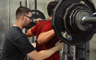 A FIT Trainer helping a patient perform barbell squat.