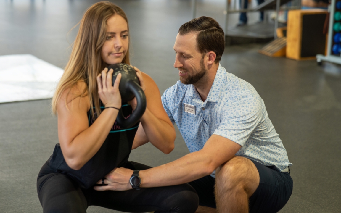 Feature image of a woman holding a kettlebell as she squats with a physical therapist holding her hip in alignment.